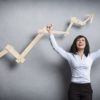 Concept: Success in business or career. Enthusiastic businesswoman with raised arms cheering in front of positive business graph, isolated on grey background.