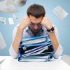 business, people, deadline, stress and paperwork concept - sad businessman with stack of folders and falling papers over blue background